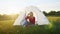 a young woman happily set up a tent on the mountain hills during sunset, for rest after a hike. woman drinking hot tea