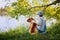 Young woman in a hat with dog Shar Pei sitting in the field and looking to the river in golden sunset light