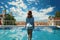 Young woman in hat and shirt standing near swimming pool with city view, A tourist woman rear view in a swimming pool enjoys the