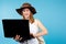 Young woman in hat smiling uses laptop on blue background with blank side space.