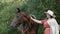 Young woman in hat stroking brown horse while grazing on farming meadow. Happy woman touching horse on forest walk at