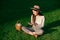 Young woman in hat uses mobile phone while sitting on green lawn at summer morning.