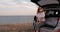 Young Woman Having Fun on Road Trip, Sitting in the Openned Trunk of Suv Car with Sea View at the Edge of Land