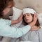 Young woman with high temperature in bed with closed eyes, her mother making compress to low the temperature.