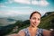 Young woman hiker smiling and taking selfie or photo with smart phone at mountain peak with mountain landscape background