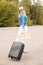 Young woman holding cardboard with inscription anywhere, black suitcase near forest, walking along road, looking back.