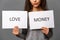 Young woman holding paper sheets with words LOVE and MONEY on grey background. Marriage of convenience