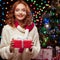 Young woman holding red gift over christmas tree and lights on b