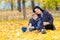 Young woman hugging teenage boy in park sitting on the ground covered with bright yellow leaves in autumn, looking at camera.