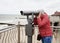 A young woman in a jacket and jeans looks through a public telescope on the waterfront and watches a beautiful panorama of the