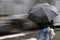 Young woman in jeans under black umbrella on a crossroad on a rainy day with traffic in motion blur,  ear view