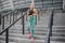 Young woman jogging down steps. She looks up to right. Well-shaped woman wear good sports uniform. She is jogging alone.