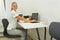 Young woman in jogging pants and t shirt sitting at her desk smiling, laptop, notepad, pencils near - physiotherapist or sport