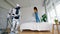 Young woman jumps on bed while a robot cleans floor.