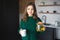 Young woman in kitchen during quarantine. Positive cheerful girl hold teapot and white cup. Posing on camera with smile.