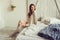 Young woman in knitted cardigan and warm socks wake up in the morning in cozy scandinavian bedroom and sitting on bed with white b