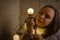 Young woman in knitted sweater wrapped in garland of glowing bulbs sitting on floor in room. Female looking at lightbulb