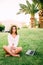 Young woman with laptop sitting on green grass with palms trees on background