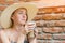 Young woman in large straw hat holding cocktail and stares in surprise at the glass. Girl with drink on brick wall background