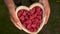 Young woman lifting a heart shaped wooden bowl of fresh red raspberries to camera
