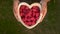 Young woman lifting a heart shaped wooden bowl of fresh red raspberries to camera and choosing one of the berries