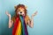 Young woman in lion mask with rainbow pride lgbt flag in mouth with raised hands and shrugging shoulders, confused expression