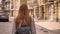 Young woman with long ginger hair walking on city street and looking back, holding backpack, urban street background