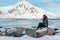 A young woman with long hair admiring the winter view of the fjord, sits on a rock near the water. On the background a large snowy