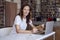 Young woman with long hair working on laptop at co-working office or library, bookshelf behind. Hold book in hands. Businesswoman