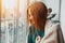 Young woman with long red hair looking through the window at home wrapped in warm comfy blanket