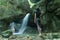 Young woman looking at splashing waterfall in rocky mountain. Caucasian Traveler standing before water stream