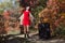 Young woman with the luggage on country road in the forest. Female person in short red dress and coat spinning arms outstretched