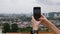 Young woman makes vertical photos of city on the smartphone standing on the mountain. Camera over the shoulder. Cloudy