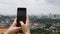 Young woman makes vertical photos of city on the smartphone standing on the mountain. Camera over the shoulder. Cloudy