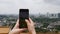 Young woman makes vertical photos of city on the smartphone standing on the mountain. Camera over the shoulder. Cloudy