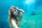 Young woman making selfie under water in the pool. In her hair is a frangipani flower. Against the background a young guy jumped i