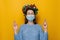 Young woman in medical face mask stands with fingers crossed, awaits miracle happen, wears spruce holiday wreath around head