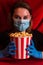 Young woman in medical mask with epidemic lettering holding bucket with popcorn on red surface with velour