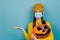 Young woman in medical mask, hold spooky pumpkin, showing empty copy space on open hand palm, wears orange sweater and hat