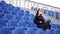 Young woman in medical mask looks away, sitting on stadium bleachers alone. Adult female in black casual clothes