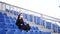 Young woman in medical mask looks away, sitting on stadium bleachers alone. Adult female in black casual clothes