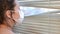 Young woman in medical mask looks out the window through the blinds