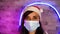 Young woman in medical mask and Santa Claus hat against illuminated wall. Close up of brunette in Christmas hat and