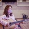 A young woman in a medical protective mask plays a home kitchen guitar. Classes over the Internet during quarantine due to
