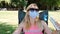 Young woman in a medical protective mask and sunglasses with funny rabbit ears hat sits in a chair and uses a fan to cool. Covid.