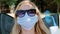 Young woman in a medical protective mask and sunglasses sits in a chair and uses a fan to cool. Covid-19 pandemic. Close-up