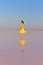 A young woman in the middle of a pink salt lake stands in a bright yellow dress and meets the sunset.
