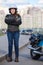 Young woman motorcyclist standing roadside near her motorcycle, full length portrait with cross arms on breast