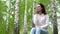 A young woman in nature is talking on the phone through headphones. A girl sits on a stump in a birch forest with
