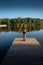 Young woman with outstretched arms standing relaxing or stretching on wooden pontoon staring at lake. evening light Back view